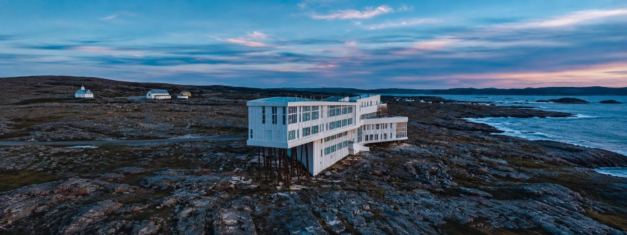 A white coastal building on stilts beside a rocky shoreline at dusk.