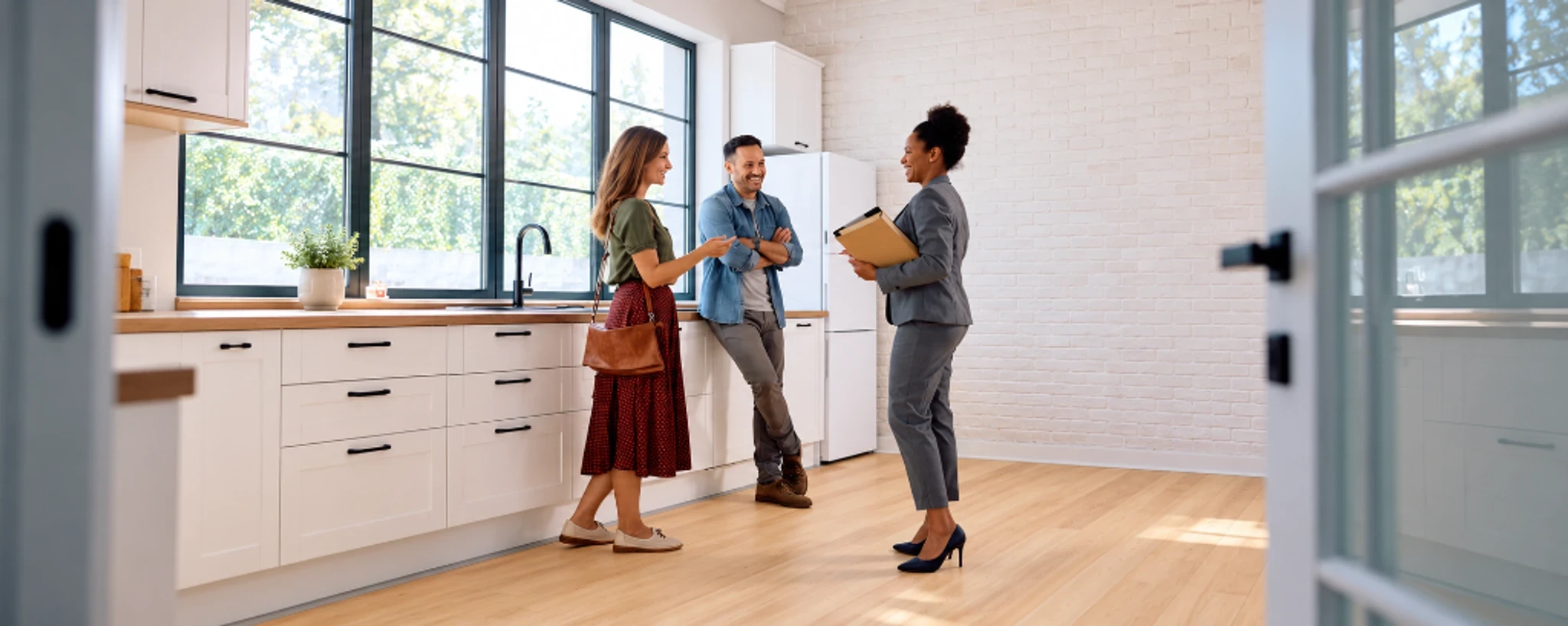 Three people talking in a bright kitchen with large windows and white cabinetry.