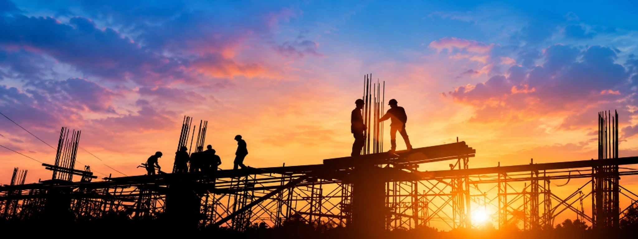 Silhouetted construction workers on scaffolding with reinforcing bars against a sunset.